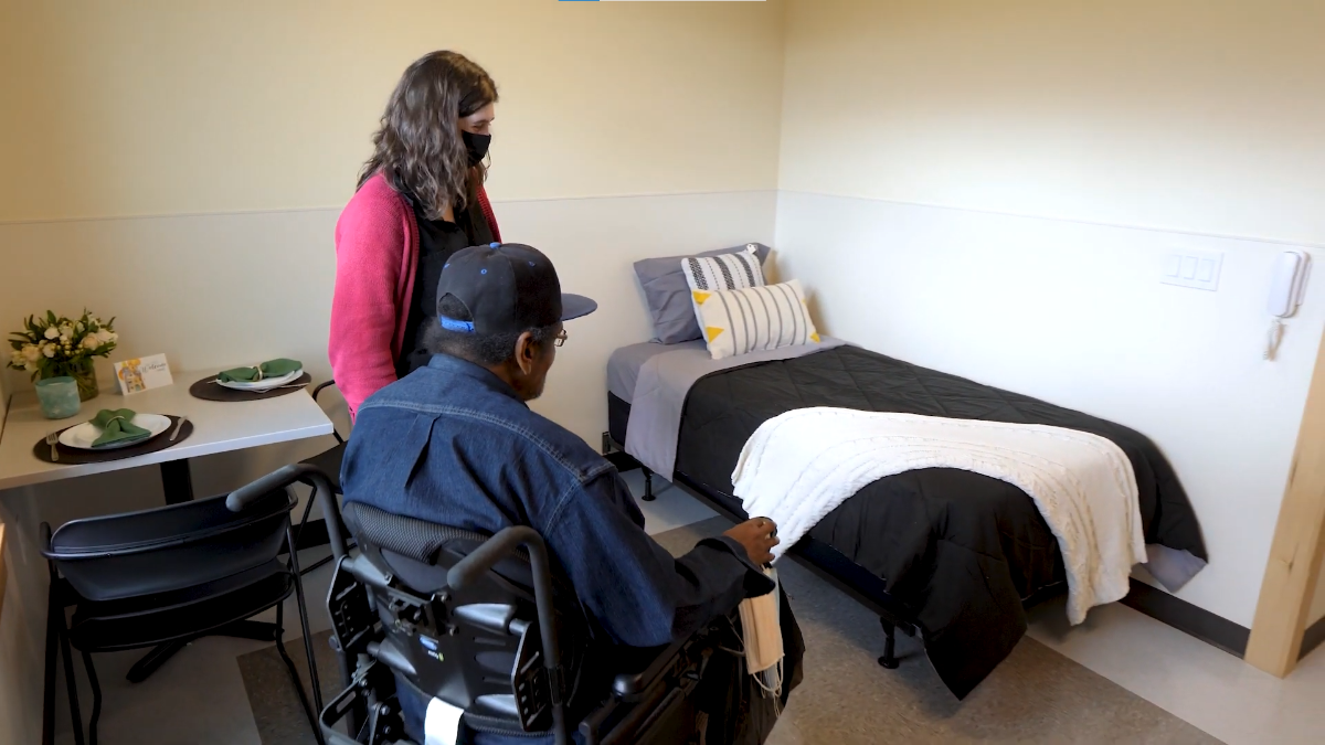 A photo of a housing resident in a wheelchair and a support service worker in front of a bed.