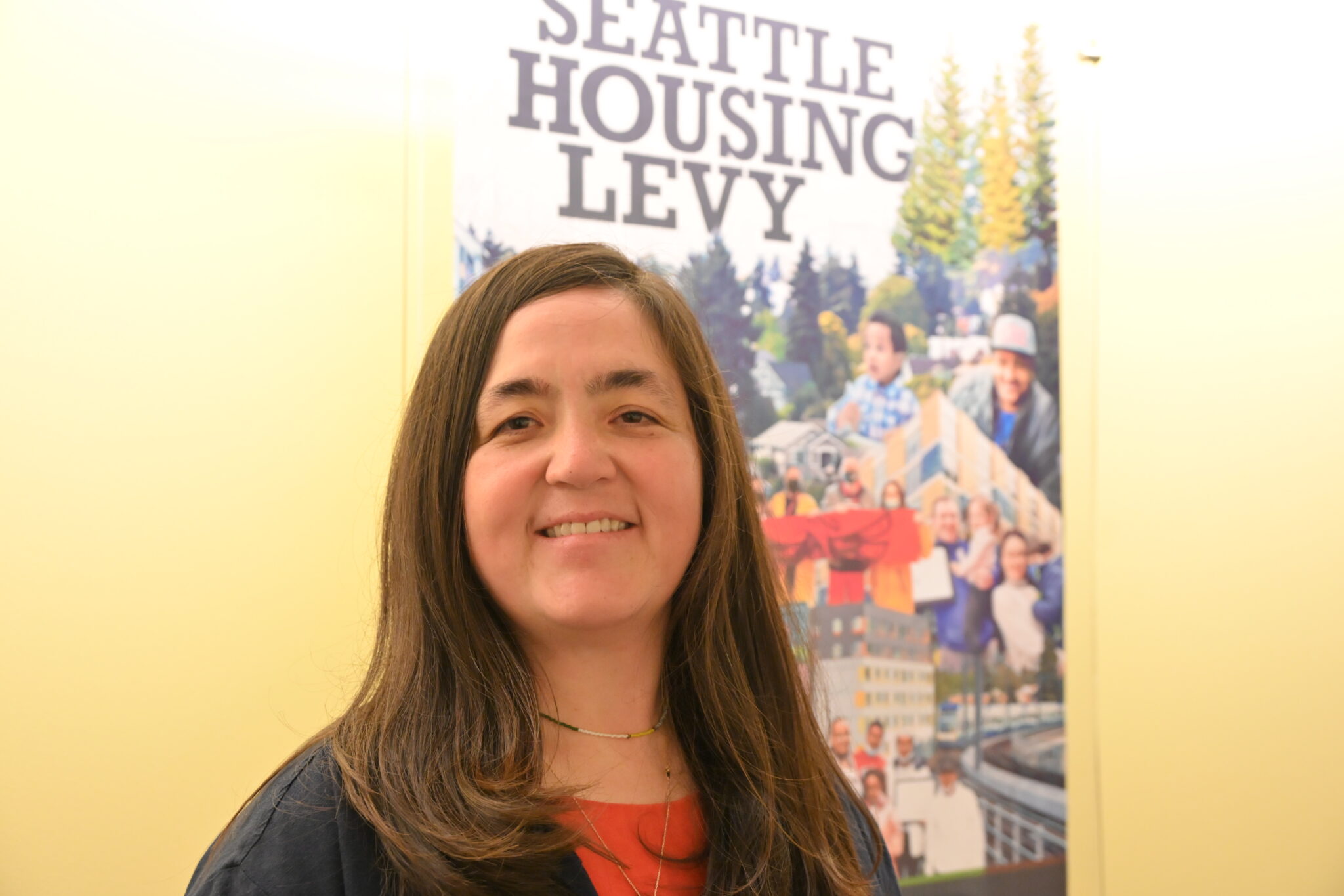 A photo of a woman standing in front of a poster with the words “Seattle Housing Levy.” She has medium long brown hair and is smiling. She is wearing an orange dress with a navy-blue blazer.